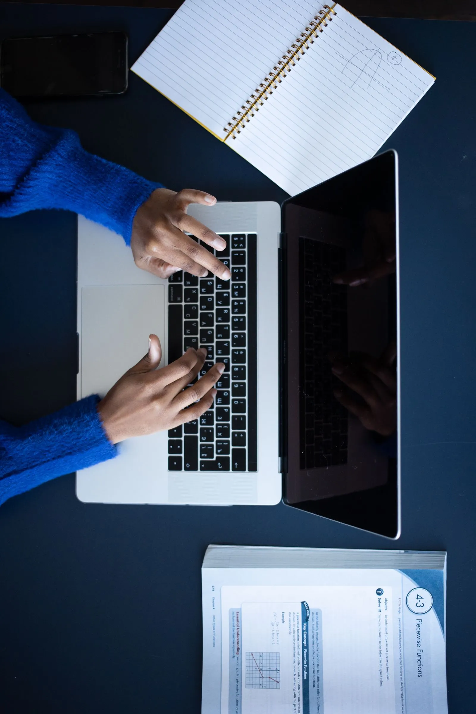 ノートパソコンのキーボードに手を置き、ノートと書類を広げた机を上から見たところ